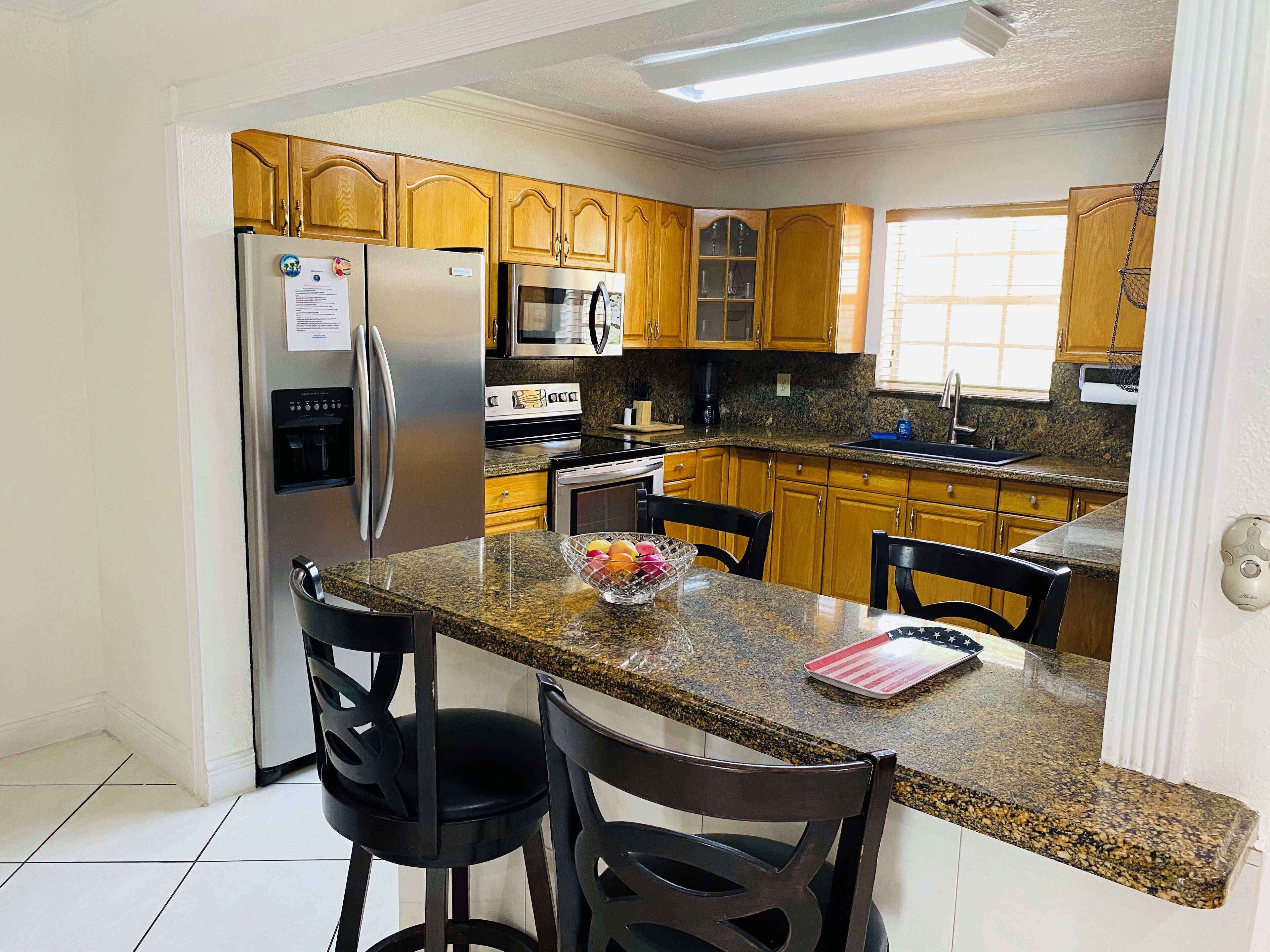 A modern kitchen with granite countertops, stainless steel appliances, wooden cabinets, and a breakfast bar with two chairs.