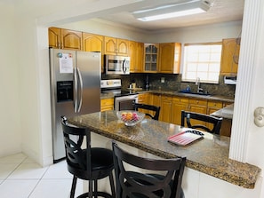 A modern kitchen with granite countertops, stainless steel appliances, wooden cabinets, and a breakfast bar with two chairs.