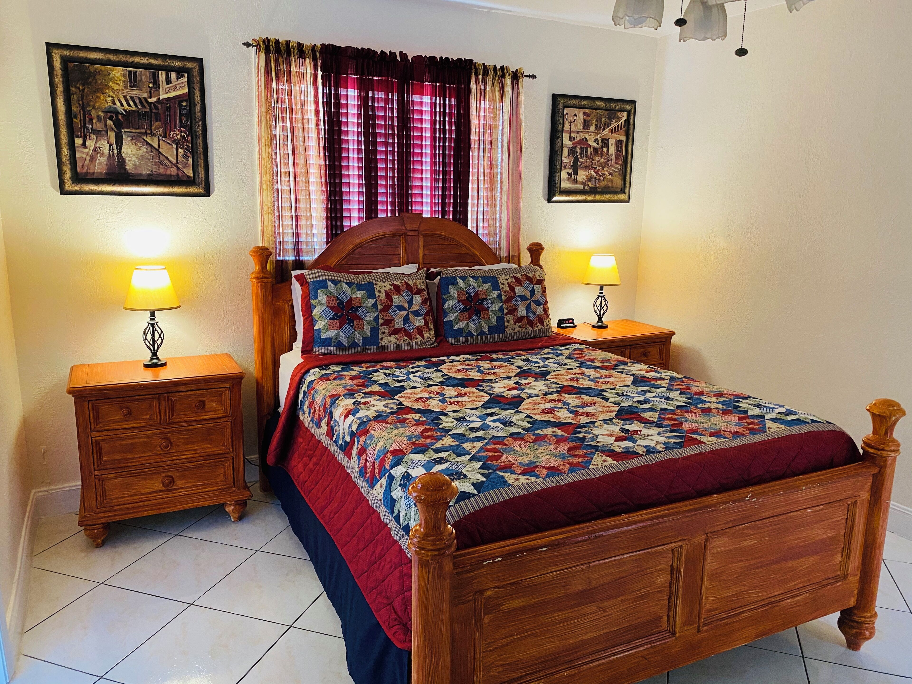 A bedroom with a wooden bed, two bedside tables with lamps, a quilt, and two framed pictures on the walls.