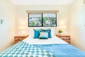 A bedroom with a bed, two bedside tables, a window, and a view of trees.