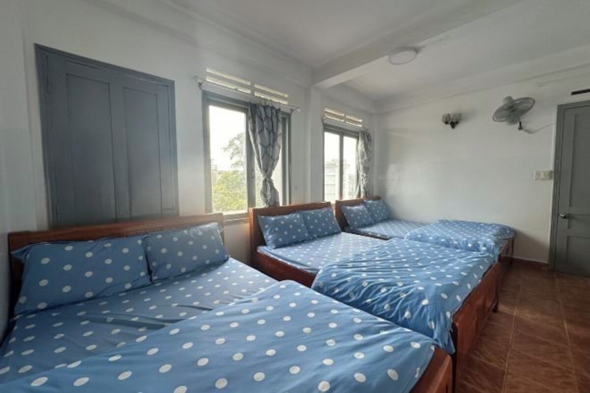 Een kamer met drie bedden, elk met blauw beddengoed en witte stippen. Er zijn twee ramen met gordijnen, een plafondventilator en een deur.