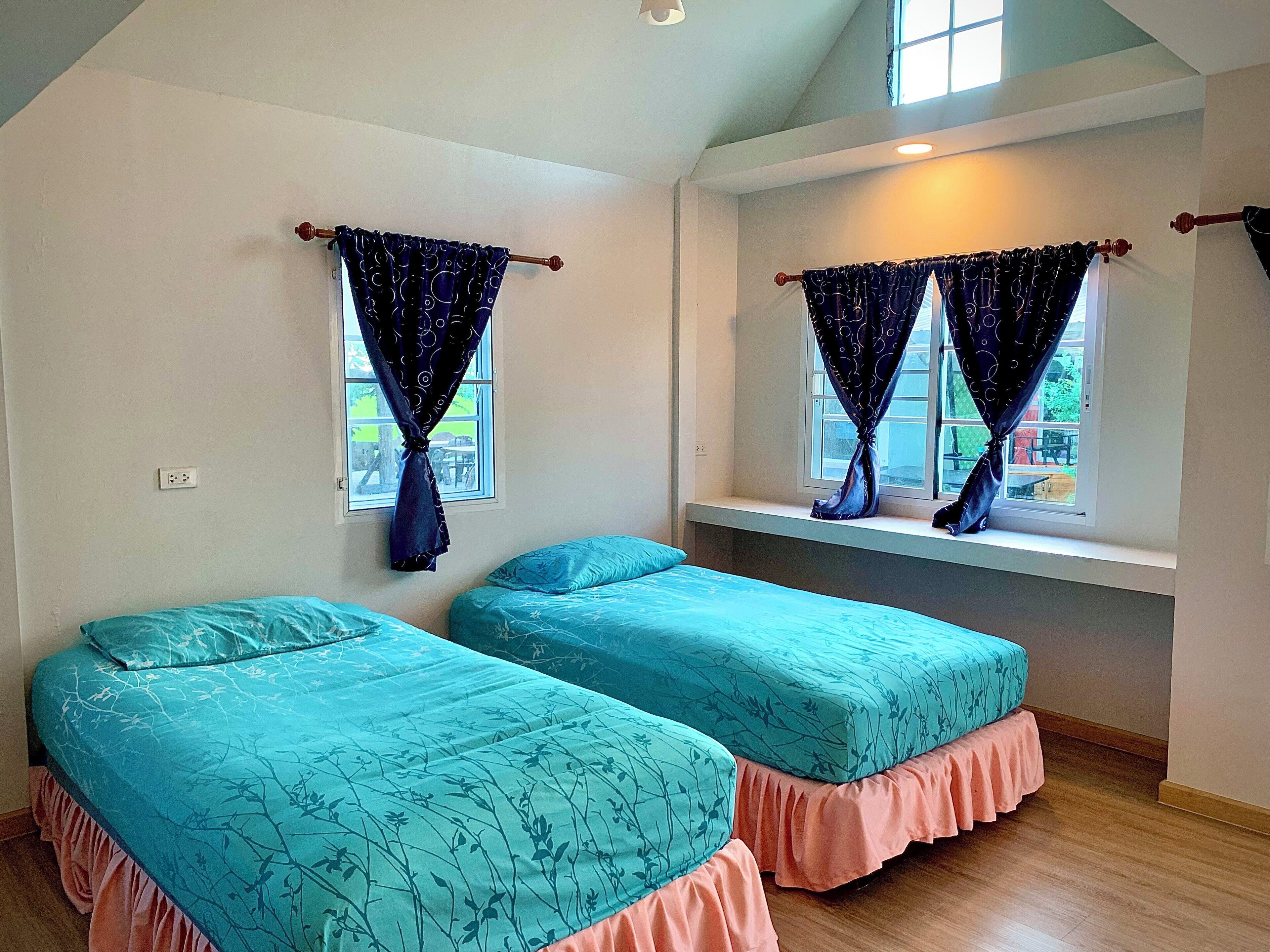 Une chambre avec deux lits, chacun recouvert d’une courtepointe à motifs floraux bleus et doté d’un couvre-lit rose à volants. Il y a des fenêtres avec des rideaux, et un petit banc près de la fenêtre au-dessus des lits.