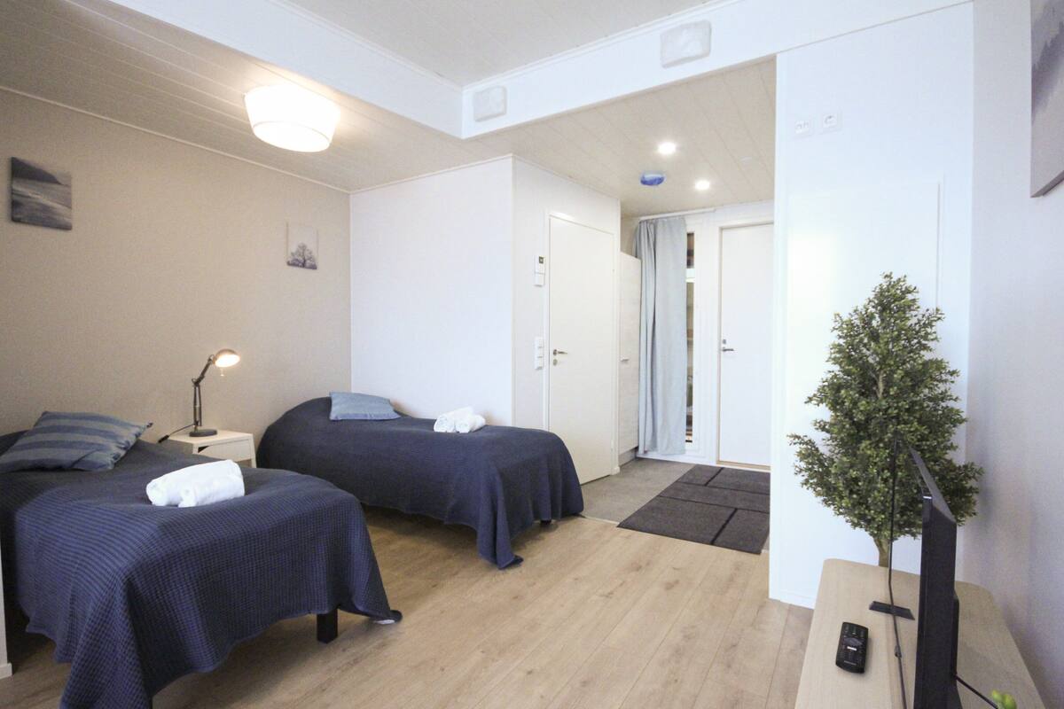 A hotel room with two beds, a small desk, a lamp, a television, and a potted plant.
