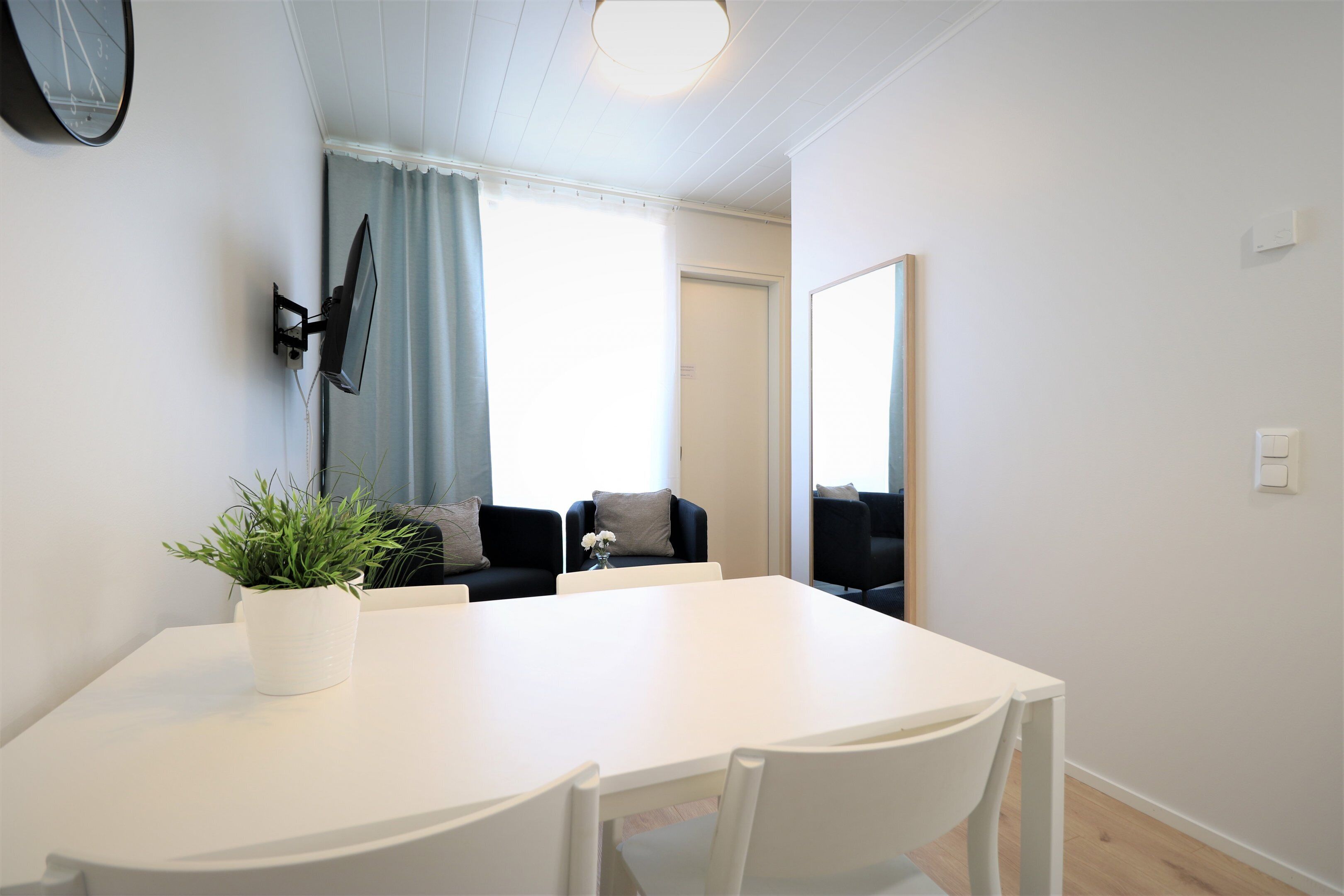Quarto moderno e bem iluminado com uma mesa de jantar branca, duas cadeiras, uma planta em vaso, uma TV de tela plana na parede e um espelho grande.