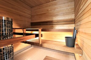 A sauna with wooden walls and benches, a stone wall section, and a black bucket with a stick.
