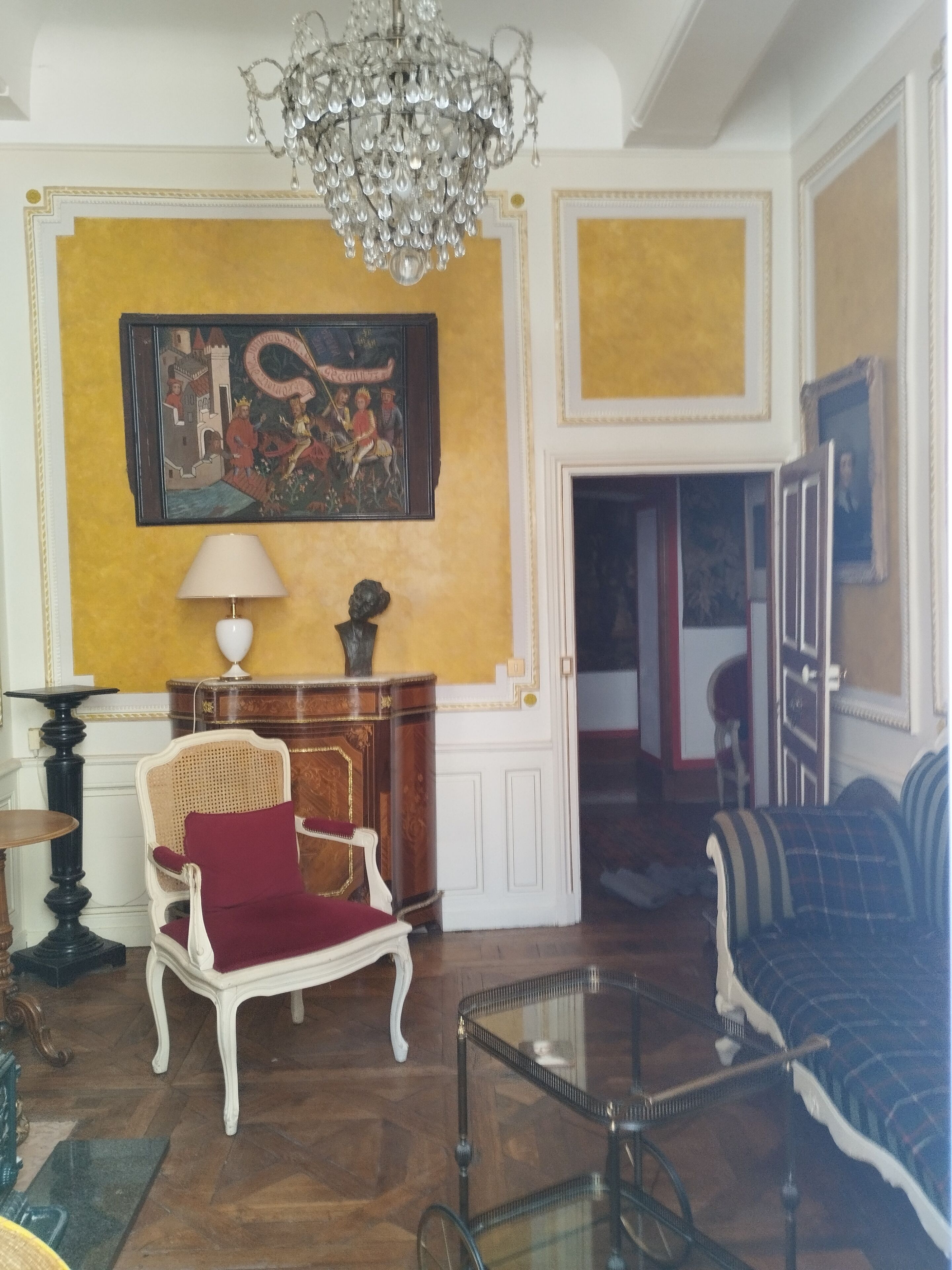 Een kamer met een kroonluchter, een schilderij, een rode stoel, een bank en een glazen salontafel.