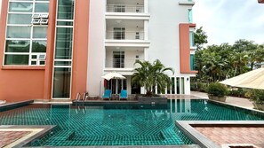 A swimming pool with a tiled deck, a lounge area with chairs and umbrellas, and a building with multiple balconies.