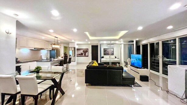 Una cocina moderna con armarios blancos, un comedor con mesa y sillas de vidrio, una sala de estar con sofá negro y televisor, y un dormitorio con cama y ventana.