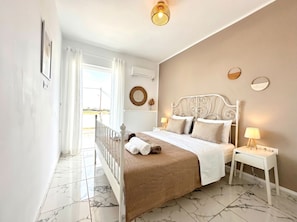 A bedroom with a bed, two bedside tables, a hanging light fixture, a window with curtains, and a view outside.