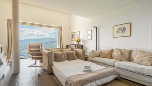A hotel room with a bed, a sofa, a chair, and a large window offering a view of the sea.