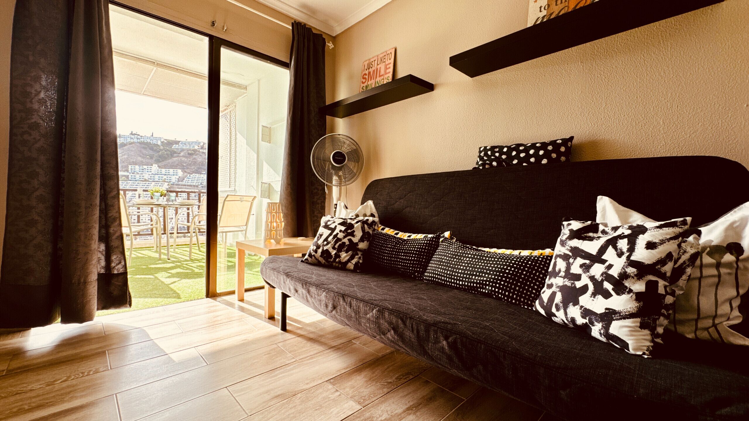 A modern living room with a dark grey sofa, patterned cushions, a standing fan, and a view of a balcony with outdoor furniture.