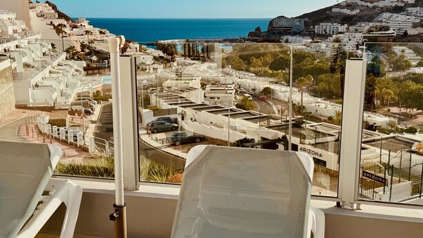 En utsikt från en balkong med vita stolar av nätmaterial, en pool och en stadsbild i bakgrunden, med byggnader och en strand.