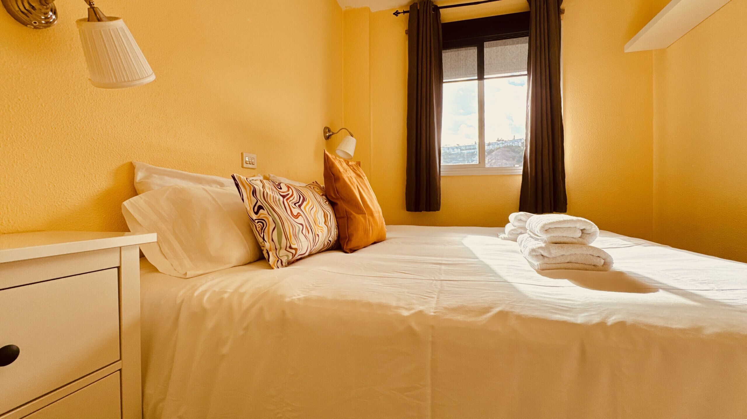 Une chambre d’hôtel avec un lit, des oreillers, une table de chevet, une lampe, une fenêtre avec des rideaux et une vue sur la ville.