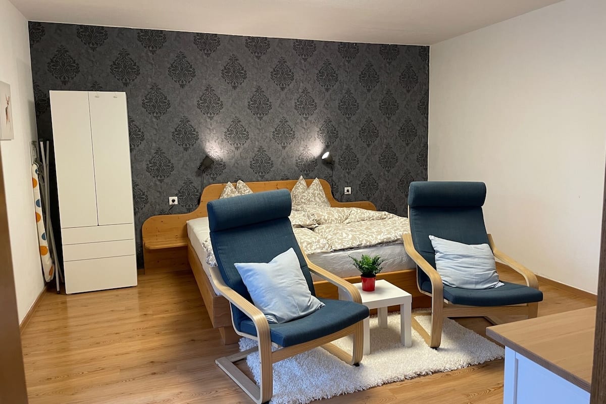 Ein modernes Schlafzimmer mit einem Bett, zwei Sesseln, einem kleinen Tisch und einem weißen Kleiderschrank.