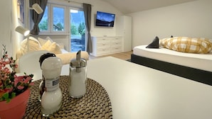 A modern hotel room with a bed, a TV, a desk with a lamp, a window with a view of a river, and a small table with a plant and candles. - Haus Sabine (Leutasch)