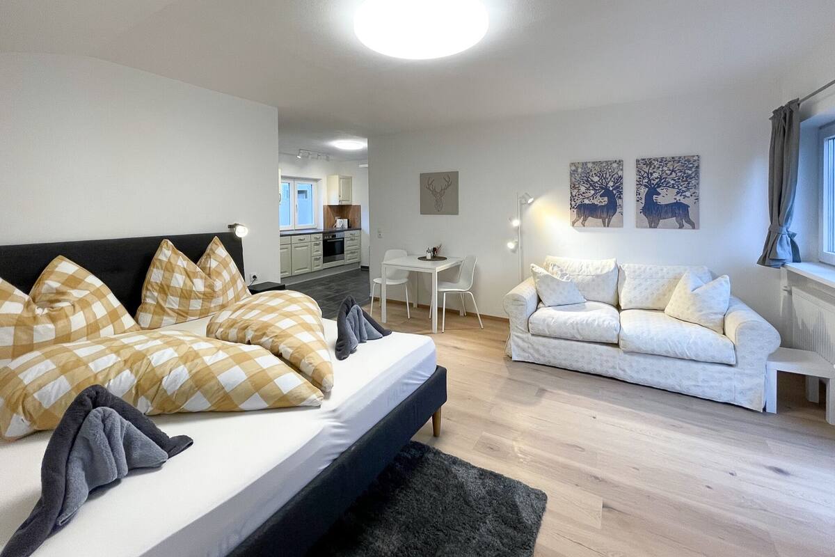 Ein modernes Schlafzimmer mit einem Bett, einem weißen Sofa, einem Schreibtisch und einer Küche im Hintergrund.