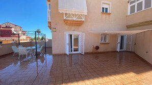 Un amplio patio exterior con suelo de baldosas, una zona de estar cubierta con sillas y una puerta corrediza de vidrio.