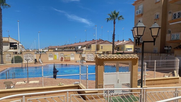 Una piscina con zona vallada, espacio de descanso cubierto y vista a edificios residenciales al fondo.