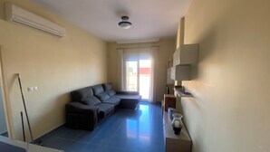 Sala de estar con sofá modular, ventilador de techo y zona de cocina con armarios y encimera.