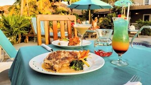 A dining table with a plate of food, a drink, and a chair in a tropical outdoor setting. - Seashell Suite, Garden & Pool Views @SapphireBeach (San Pedro Town)