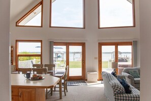 A spacious living area with a dining table, chairs, and a sofa. There are large windows providing a view of the outdoors.