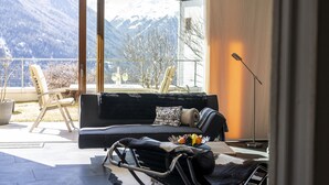 Ein modernes Wohnzimmer mit einer Couch, einem Esstisch mit Stühlen, einem kleinen Tisch mit Obstschale und Blick auf schneebedeckte Berge durch große Fenster.