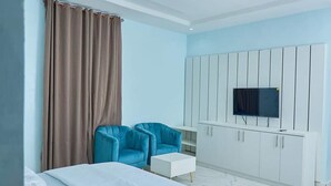 Een moderne hotelkamer met een bed, twee blauwe fauteuils, een klein wit tafeltje, een televisie bevestigd aan een wit kastje en een groot raam met gordijnen.