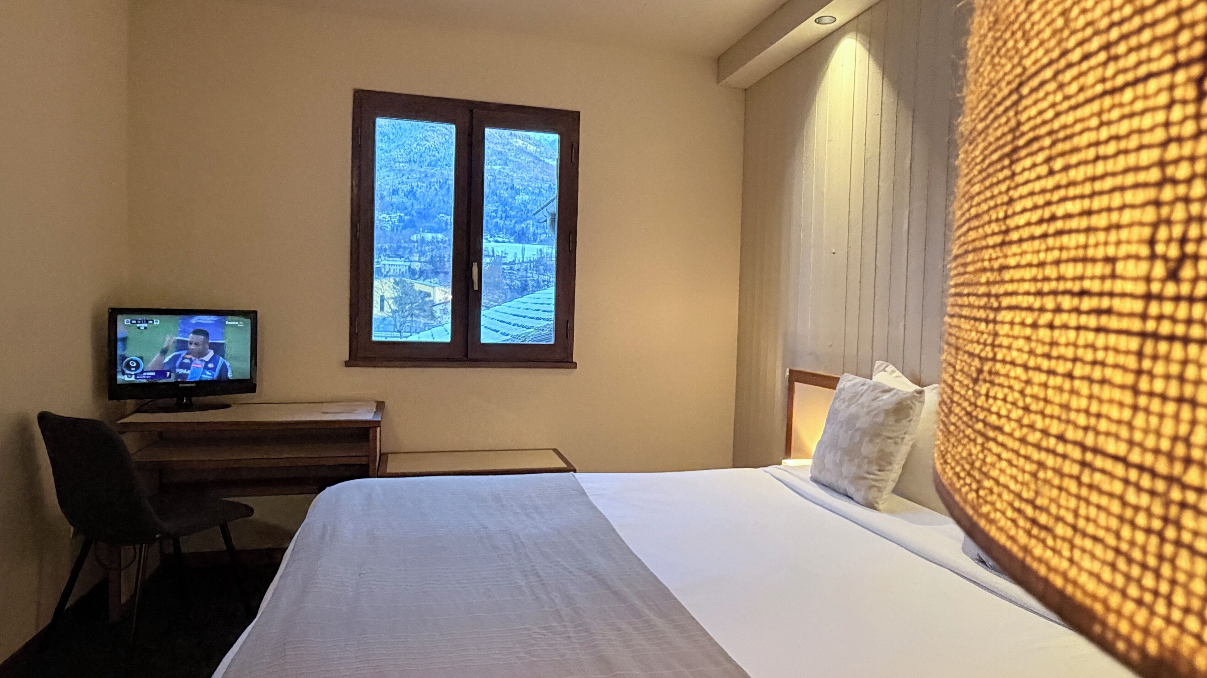 A hotel room with a bed, a desk with a television, and a window with a view of a snowy landscape.