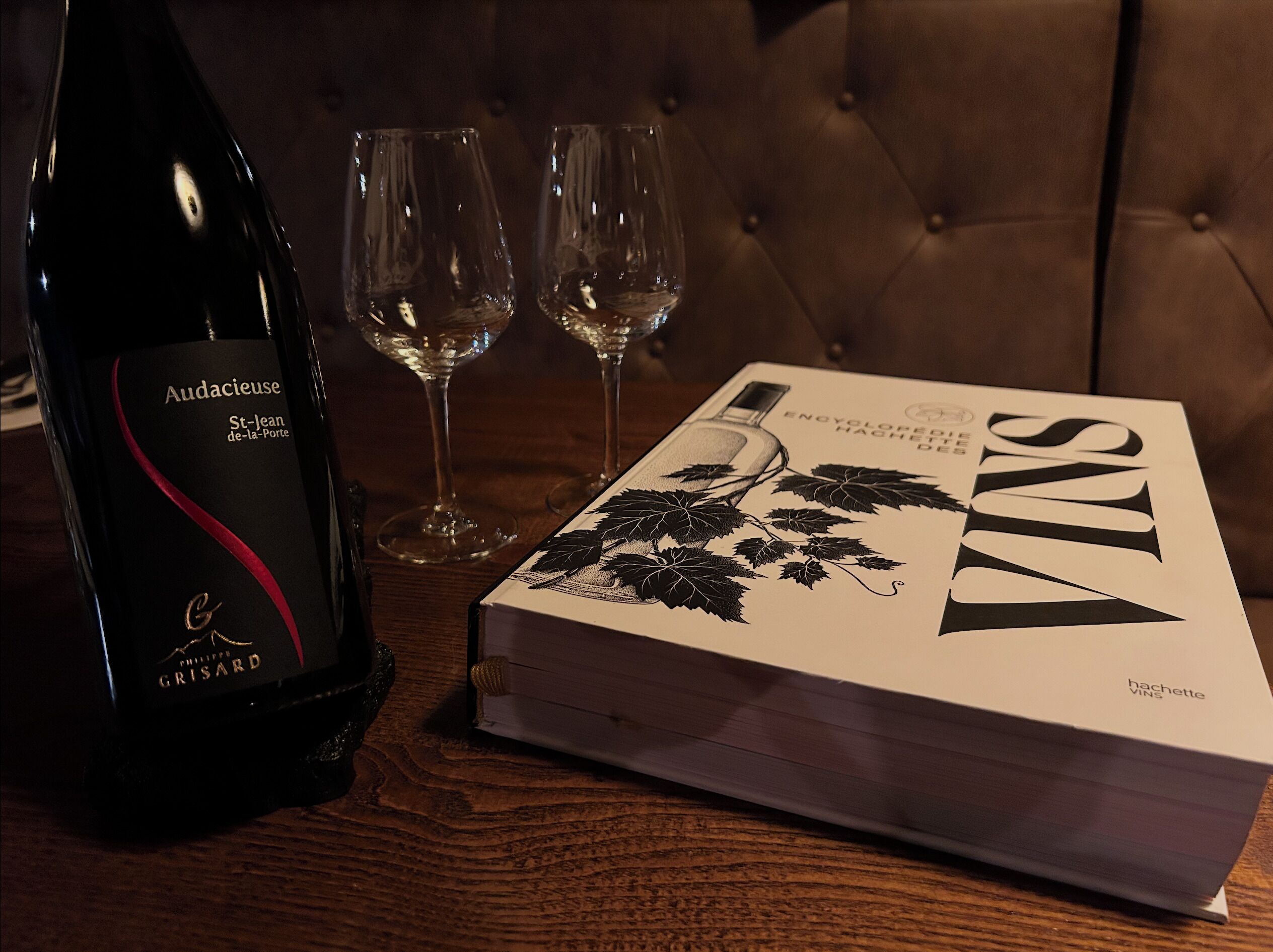 A bottle of Audacieuse St-lean de-la-Porte wine, two empty wine glasses, and a book titled 'VINs' on a wooden table.