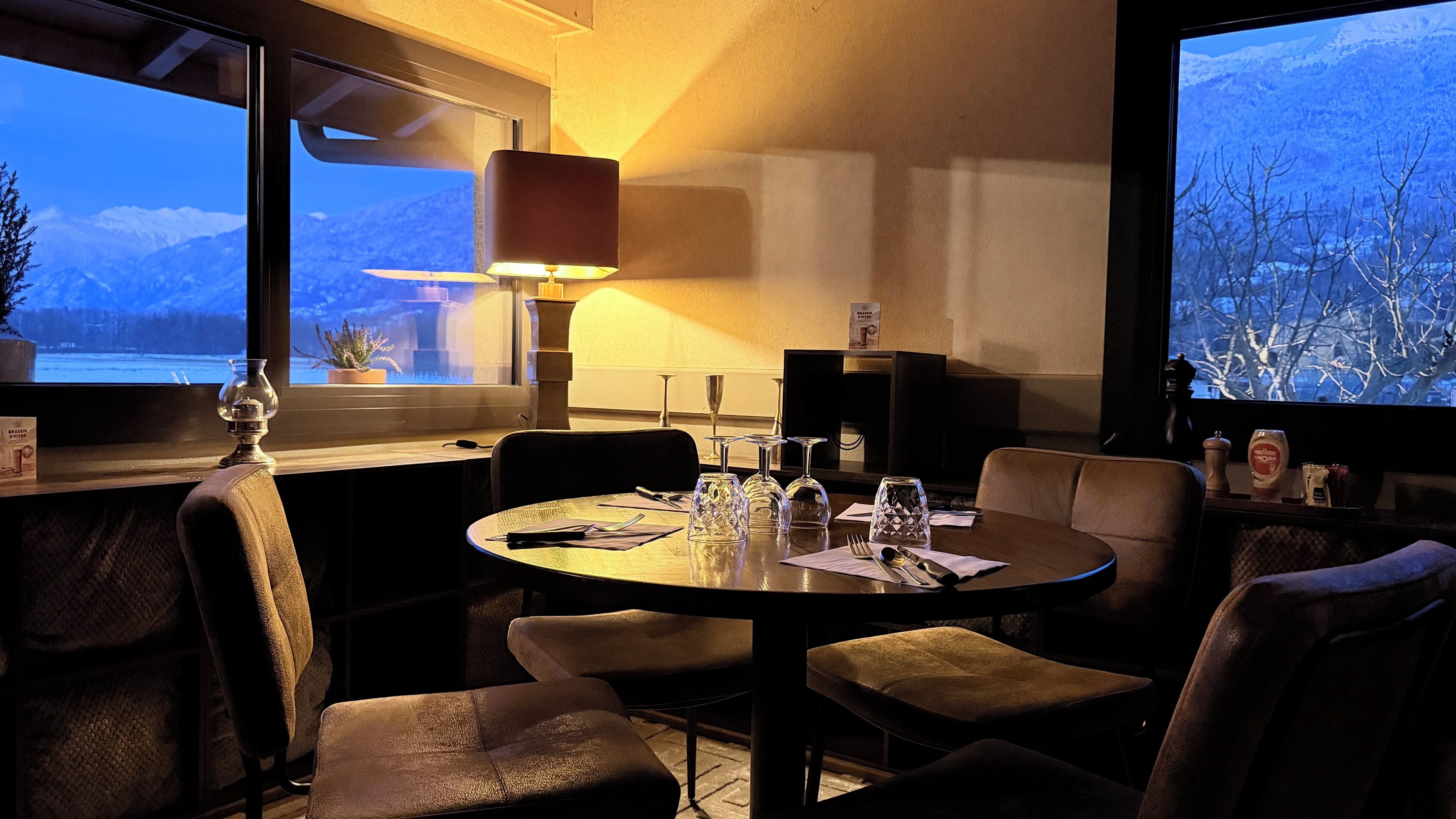 Een eetgedeelte met een ronde tafel, stoelen, een lamp en uitzicht op bergen door het raam.