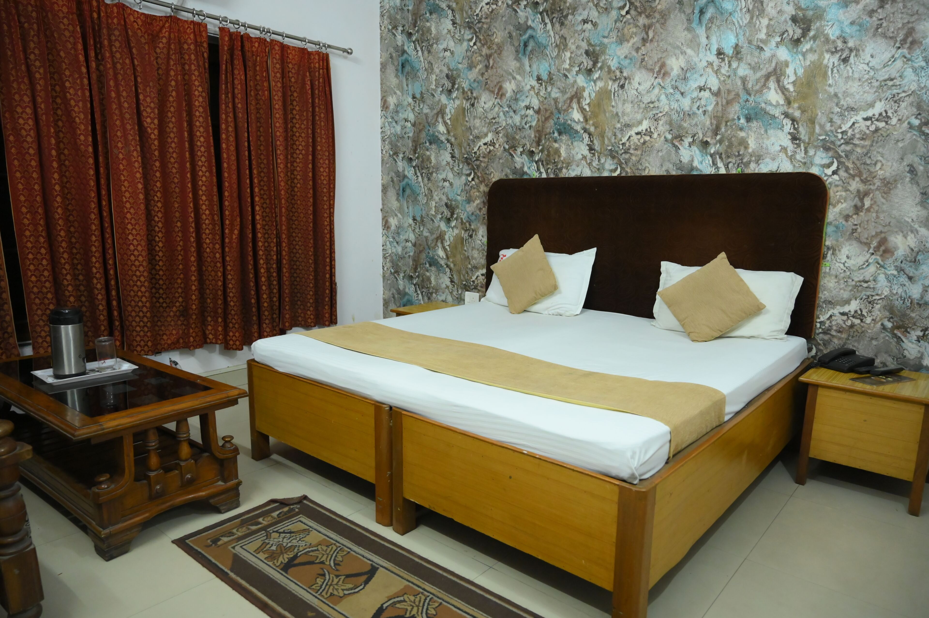 Un dormitorio con una cama de madera, una mesita de noche con un teléfono, una mesita de noche con una lámpara y una alfombra estampada.