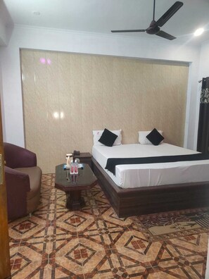 A hotel room with a bed, a chair, a small table with water bottles, a ceiling fan, and patterned floor tiles.