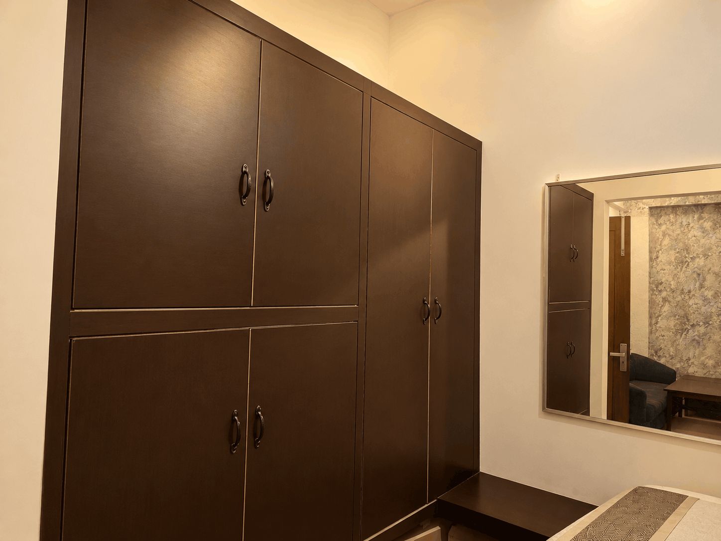 A room with a large wardrobe, a small table, and a mirror reflecting a sofa and a patterned wall.