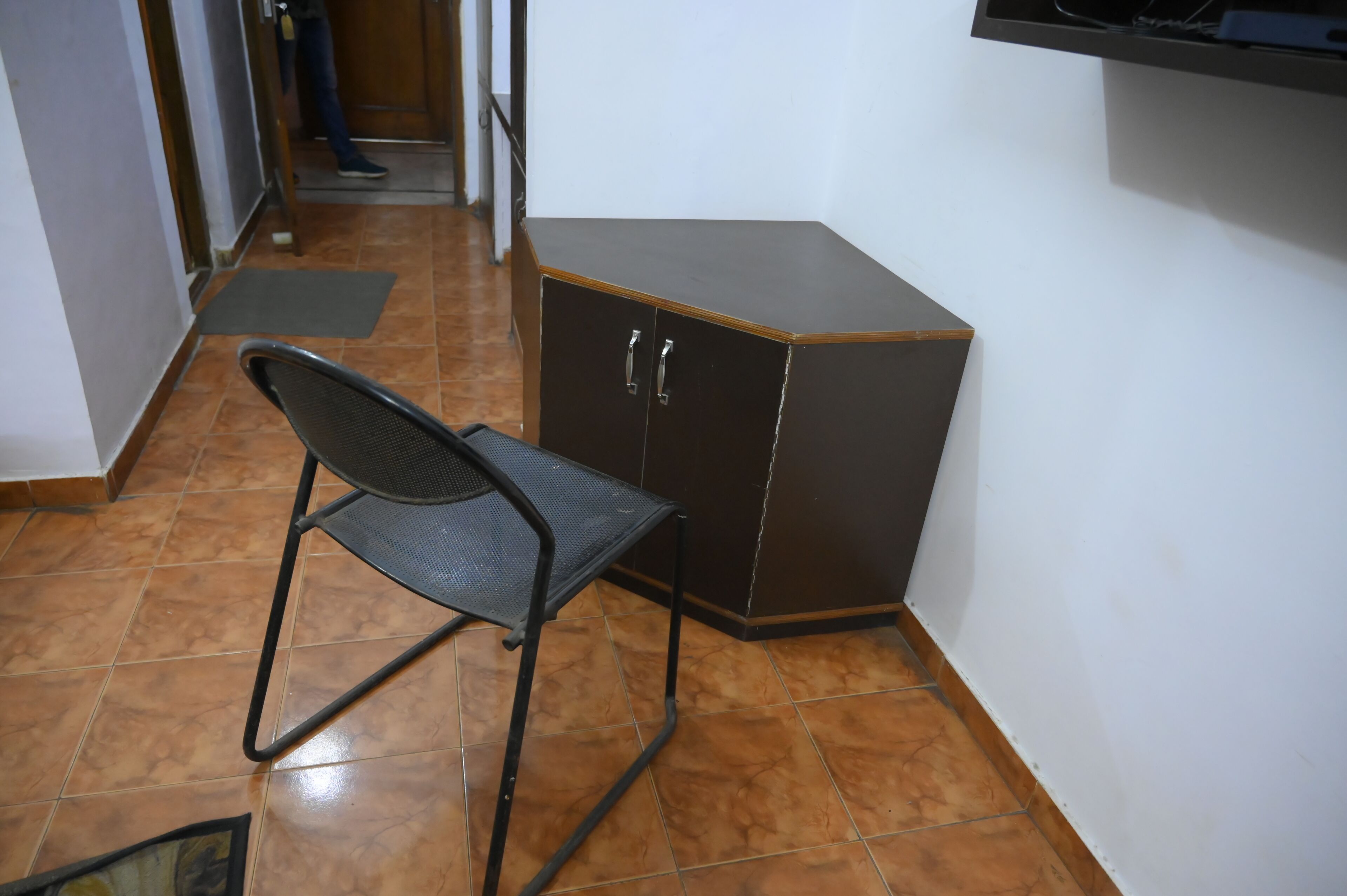 Een kleine kamer met een metalen stoel, een donker houten kast en een betegelde vloer.