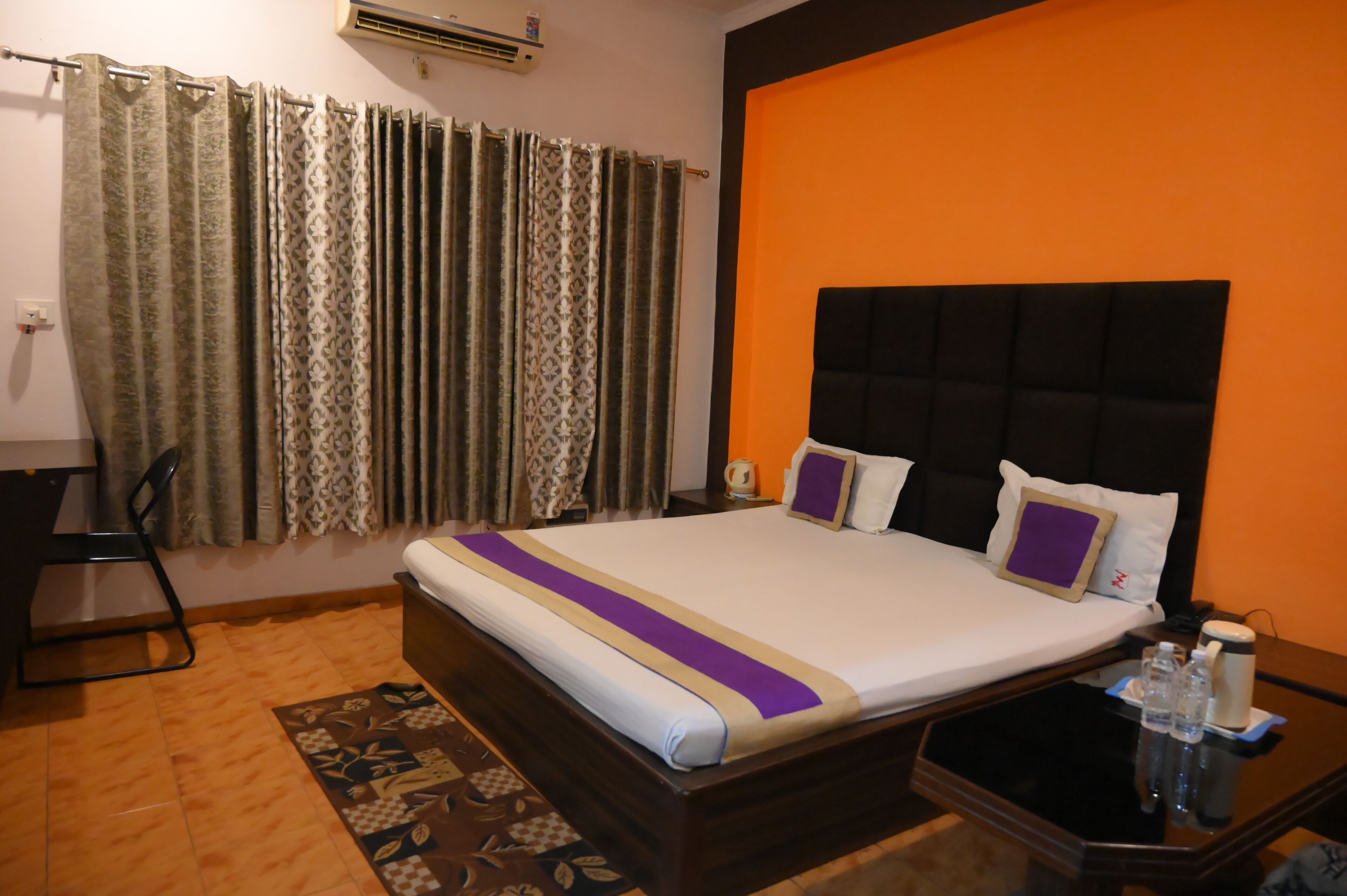 A hotel room with a bed, a nightstand, a chair, a window with curtains, and an air conditioning unit.
