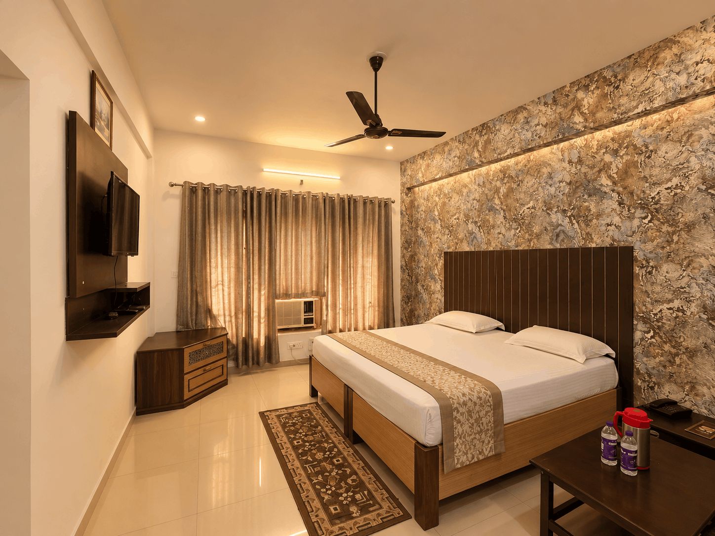 A hotel room with a bed, a TV, a ceiling fan, a wall-mounted air conditioner, and a nightstand with a phone and bottles.