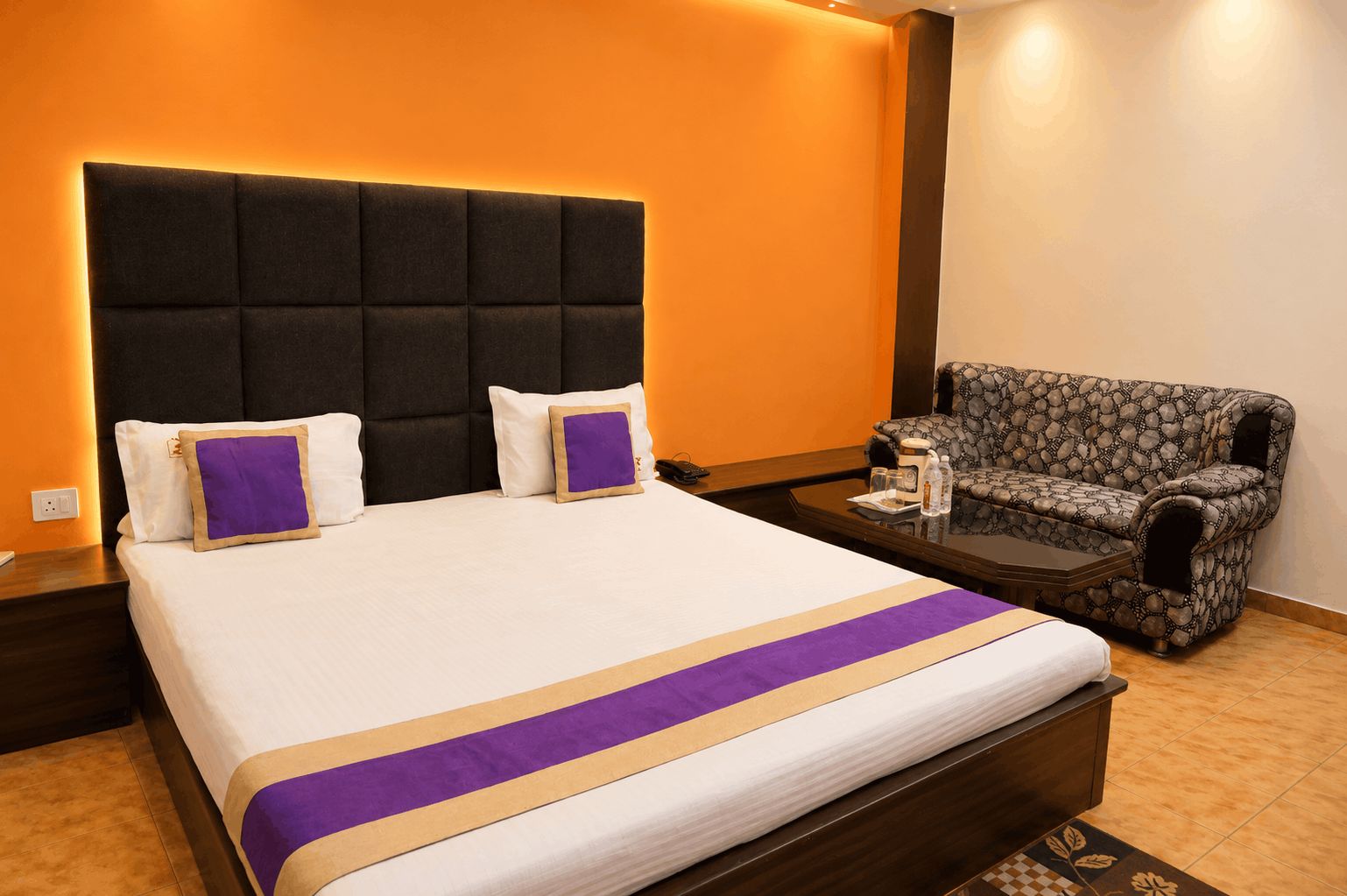 Habitación de hotel con una cama grande, cabecera con iluminación integrada, un sillón estampado y una mesita con una botella y vasos.