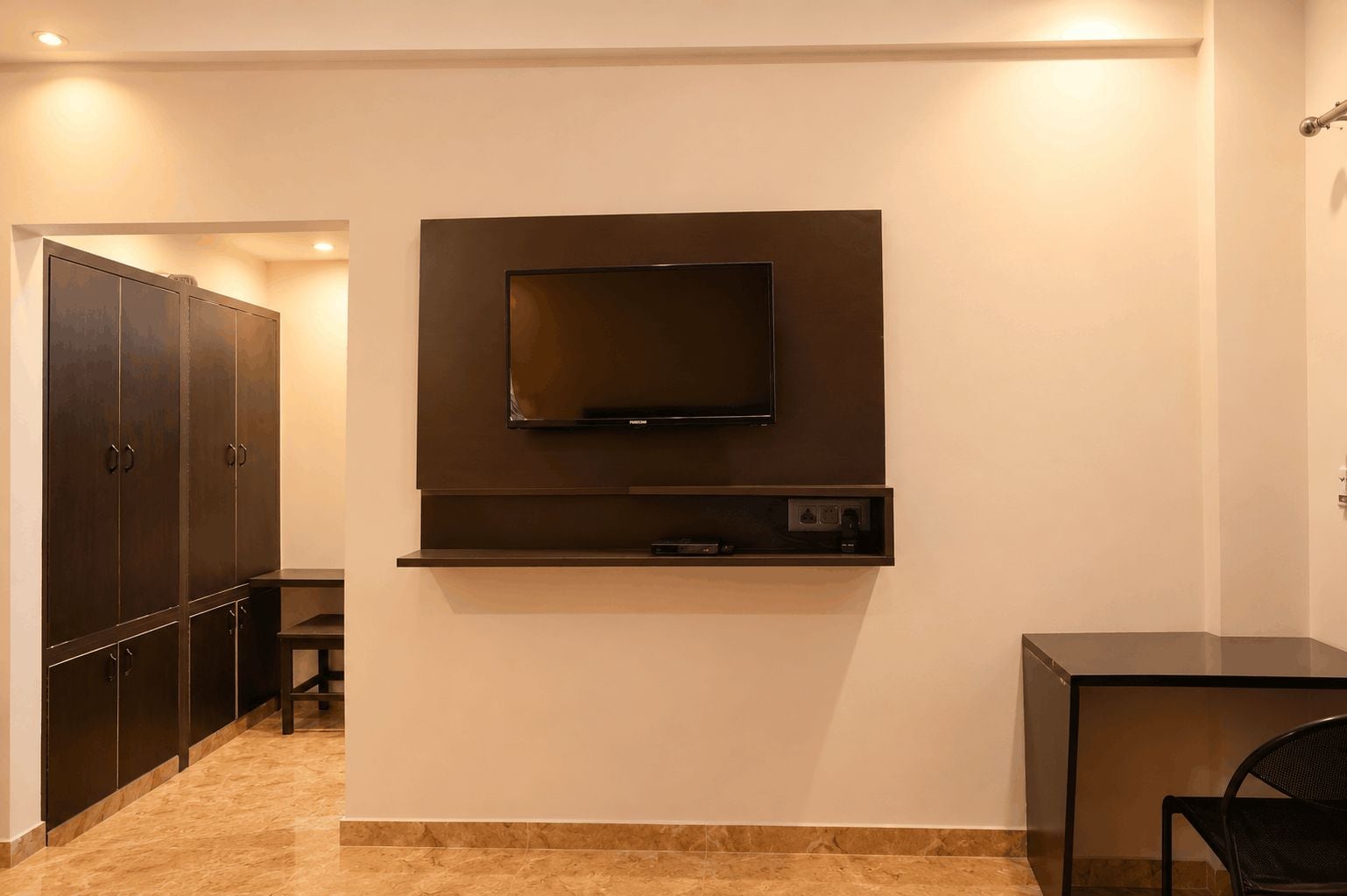 A room with a flat-screen TV mounted on the wall, a small shelf below it, and a chair to the right.