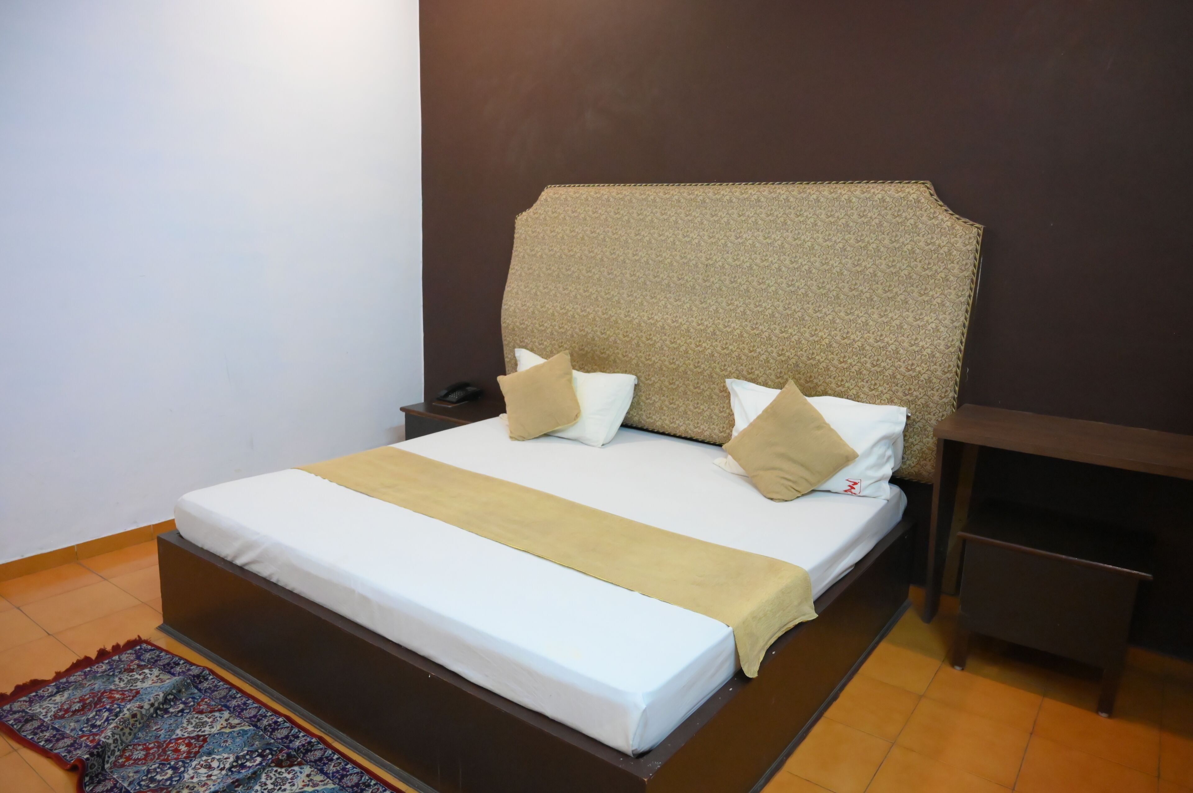 A neatly made bed with a headboard, a bedside table, and a patterned rug on the floor.