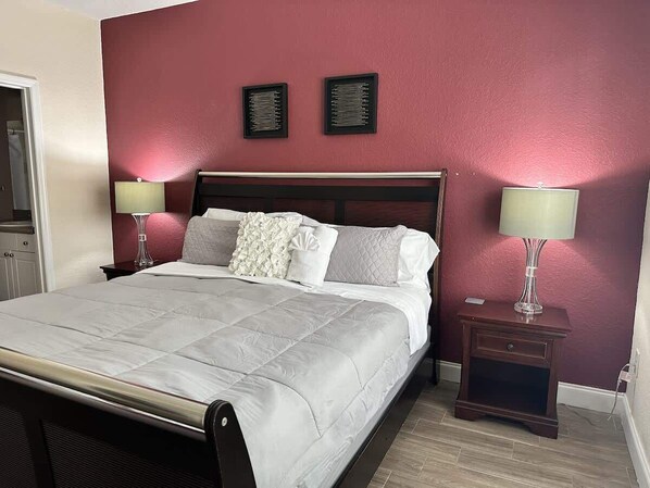 A neatly made bed with a white duvet and pillows, a dark wooden headboard, two bedside tables with lamps, and a red wall with two framed pictures.