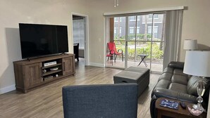 A living room with a flat-screen TV, wooden entertainment unit, grey sofa, and a red chair near a sliding glass door leading to a balcony. - Crescent Palms - Near Brandon, Tampa, Riverview (Tampa)