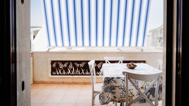 A balcony with a table and chairs, a view of buildings, and a striped curtain. - Appartamento a Torre Canne con Balcone (Fasano)