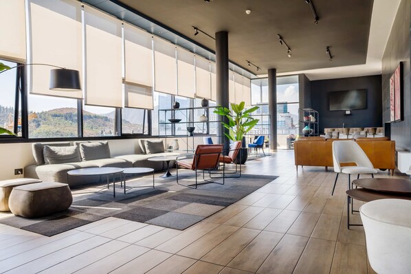 A spacious living area with a sofa set, coffee table, and chairs, large windows with blinds, and a flat-screen TV mounted on the wall.