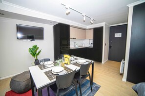Un espace repas moderne avec une table blanche dressée pour quatre personnes, des chaises noires, un téléviseur à écran plat fixé au mur et une cuisine en arrière-plan.
