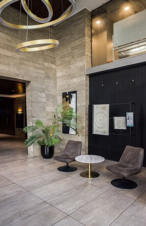 Un hall d’entrée au design moderne, comprenant un espace salon avec deux fauteuils et une table, une plante en pot et un panneau d’information fixé au mur.