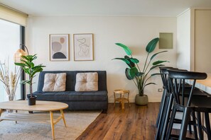 A modern living room with a dark grey sofa, two cushions, a round wooden table, a small round wicker stool, and a large potted plant.