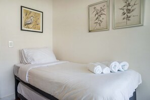 Una cama individual con ropa de cama blanca y toallas enrolladas, un interruptor de luz en la pared y tres ilustraciones botánicas enmarcadas.