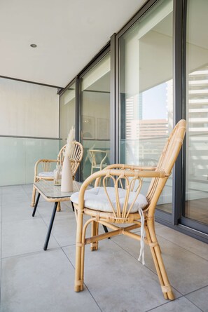 Un balcón con dos sillas de mimbre, una mesa, ventanas de vidrio grandes y vistas a edificios.