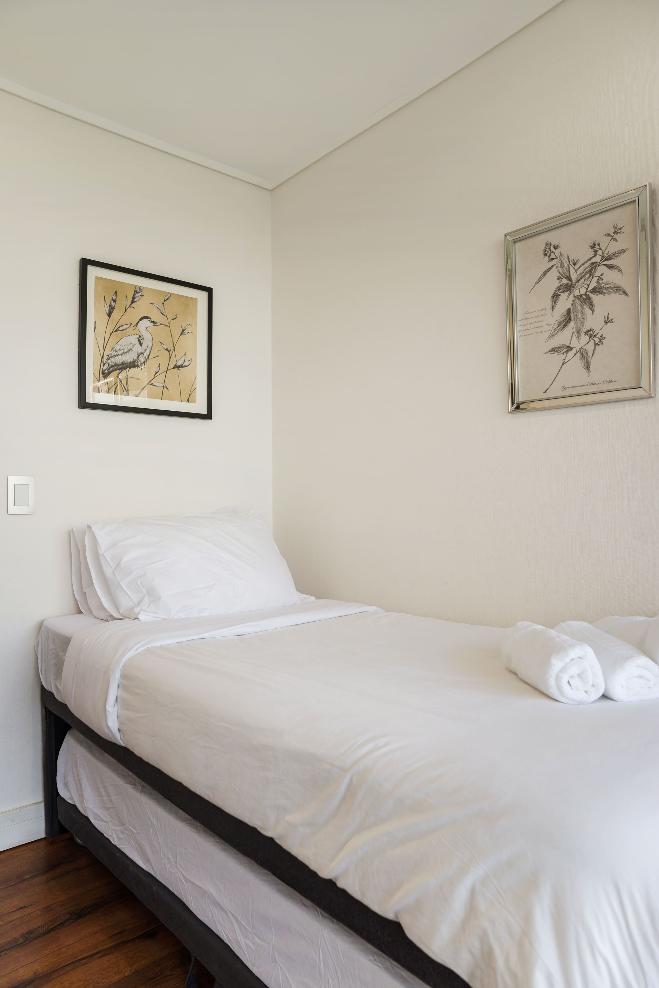 Een enkel bed met witte beddengoed, een donker hoofdbord en twee ingelijste botanische afbeeldingen aan de muur.