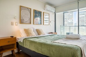 A modern bedroom with a large bed, a nightstand, a bedside table, and a window with a view of buildings. - Comfortable 2BR/2BA Apt. for 5 guests with Gym! (Las Condes)